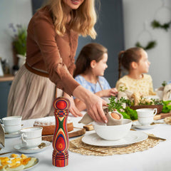 A woman sets a table with bowls and cups, adorned with greenery from Totally Bamboo's Marrakesh Collection. Three children sit eagerly at the table, where a colorful pepper grinder adds a vibrant kick to the setting, while the Baltique® Marrakesh Collection Refillable Salt Shaker promises endless flavor in this bright, festive atmosphere.