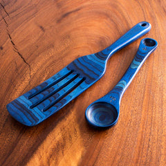 A wooden surface beautifully displays a Baltique® Malta Collection 2-Piece Utensil Set by Totally Bamboo, featuring a blue wooden spatula and a 2-in-1 Measuring Spoon, both adorned with intricate grain patterns and a sleek finish.