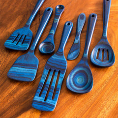 From the Baltique® Malta Collection by Totally Bamboo, this 7-piece cooking utensil set includes spatulas and spoons artistically displayed on a brown wooden surface. Crafted from colored birch, these utensils showcase distinctive striped and circular patterns in an array of blue shades.