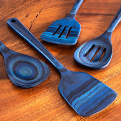 Four kitchen utensils from the Baltique® Malta Collection by Totally Bamboo are arranged on a textured wooden surface. This colored birch set includes slotted spatulas and spoons in blue and black, each featuring a unique layered pattern.