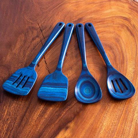 Totally Bamboo's Baltique® Malta Collection 4-Piece Cooking Utensil Set presents a delightful assortment of four blue wooden kitchen tools, including a spatula, slotted spatula, ladle, and spoon. Made from colored birch, each utensil features a unique wavy pattern in different shades of blue against the lush brown wooden surface.