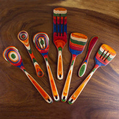 The Baltique® Marrakesh Collection 7-Piece Cooking Utensil Set by Totally Bamboo features seven vibrant wooden kitchen tools, such as spoons and spatulas. These utensils are elegantly arranged in a semicircle on a wooden surface, with their colorful birch handles displaying striking multicolored stripes and patterns.