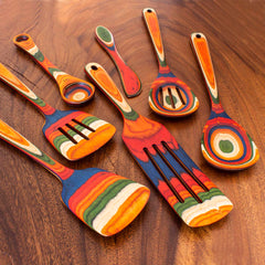 A variety of kitchen utensils from the Totally Bamboo Baltique® Marrakesh Collection 7-Piece Cooking Utensil Set are arranged on a wooden surface. Each utensil, such as spatulas and a spoon, displays vibrant, swirling patterns in shades of red, blue, orange, green, and natural wood tones made from colored birch.