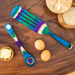 The Baltique® Mumbai Collection 2-Piece Utensil Set by Totally Bamboo features a colorful wooden spatula and measuring spoon crafted from colored birch in vibrant shades of green, blue, and purple. Displayed on a wooden table along with two eggs, butter, and granulated sugar, these utensils add a lively touch to any kitchen setting.