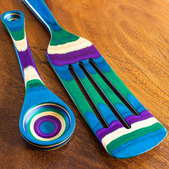 The Baltique® Mumbai Collection 2-Piece Utensil Set by Totally Bamboo showcases a vividly colored striped wooden spatula with slots and a spoon with a round bowl, featuring vibrant hues of blue, green, purple, and yellow on a wooden surface.