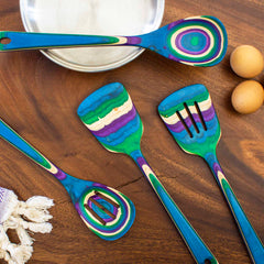 The Baltique® Mumbai Collection 4-Piece Cooking Utensil Set by Totally Bamboo showcases four vibrant cooking utensils made from colored birch, featuring lively patterns in blue, purple, green, and yellow. These utensils are displayed on a wooden surface accompanied by a metal bowl, eggs, and a fringed fabric.