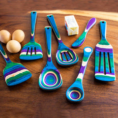 The Totally Bamboo Baltique® Mumbai Collection 7-Piece Cooking Utensil Set showcases a vibrant array of colored birch kitchen tools adorned with swirling patterns, tastefully displayed on a wooden surface. Completing this charming kitchen scene are three brown eggs and a stick of butter in its wrapper nearby.