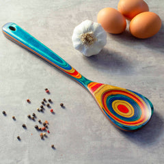 A lively Baltique® Montego Bay Collection Cooking Spoon from Totally Bamboo, measuring 12-1/2 inches, featuring concentric circles lies on a gray surface. It is surrounded by a garlic bulb, three eggs, and scattered peppercorns. This colorful birch-crafted utensil enhances the style of any kitchen environment.