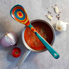 A saucepan filled with tomato soup, garnished with herbs, sits on a gray surface. A vibrant cooking spoon from the Baltique® Montego Bay Collection by Totally Bamboo rests on top. Surrounding the pan are garlic cloves, a halved onion, a whole tomato, and scattered peppercorns.