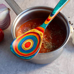 A colorful Totally Bamboo Baltique® Montego Bay Collection Cooking Spoon, 12-1/2", with a circular pattern rests on a pot of tomato soup sprinkled with herbs. Nearby, an onion, tomato, and garlic bulb sit on the gray countertop.