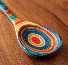 The Totally Bamboo Baltique® Montego Bay Collection Cooking Spoon, measuring 12-1/2 inches, showcases a colorful design with concentric circles in red, blue, yellow, and natural wood hues on colored birch. The handle features a similar striped pattern, adding a vibrant and artistic touch to your kitchen.