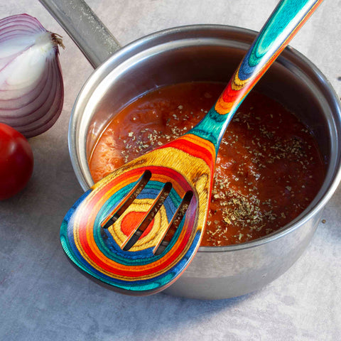 A stainless steel pot filled with tomato sauce, garnished with herbs, sits on a grey countertop. A vibrant Totally Bamboo Baltique® Montego Bay Collection Slotted Spoon, 12-1/2" rests on the pot. Beside it, a sliced red onion and a whole tomato complete the scene.