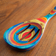 A slotted spoon from the Baltique® Montego Bay Collection by Totally Bamboo rests on a brown wooden surface. This 12-1/2" wooden utensil showcases vibrant, layered rings in shades of blue, red, orange, and yellow from colored birch, creating a rainbow-like appearance.