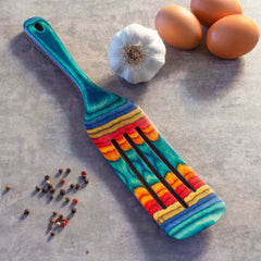 A vibrant kitchen tool, the Baltique® Montego Bay Collection Spurtle by Totally Bamboo, measuring 13 inches, displays its striking blue, yellow, and red striped design on a stone countertop. Nearby are three brown eggs, a garlic bulb, and some scattered peppercorns—perfectly setting the stage for culinary adventures.