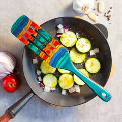 A vibrant Baltique® Montego Bay Collection Spurtle from Totally Bamboo rests on a frying pan filled with sliced zucchini, diced onions, and seasoning. Surrounding the pan are garlic cloves, a red onion, and a tomato on a light gray surface.