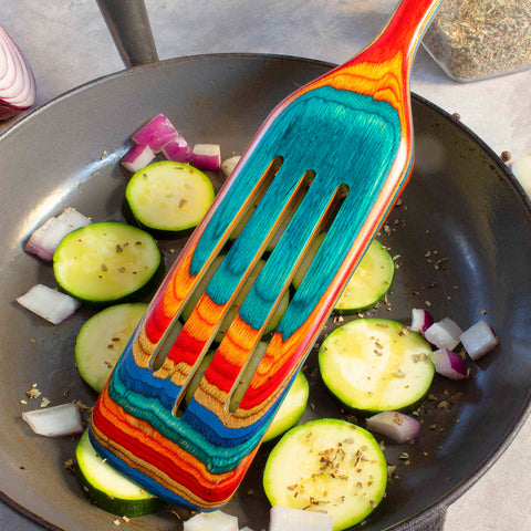 A vibrant, multicolored Baltique® Montego Bay Collection spurtle by Totally Bamboo rests on a pan filled with sliced zucchini and chopped red onion. Seasoning is sprinkled on top, creating a lively kitchen environment.