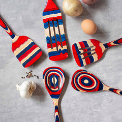 These five wooden kitchen utensils from the Baltique® Old Glory Collection by Totally Bamboo, including a 12-1/2" slotted spoon, showcase colorful red, blue, and natural wood patterns in vibrant colored birch. They are arranged in a circle on a gray surface, surrounded by ingredients such as garlic bulbs, eggs, and potatoes.