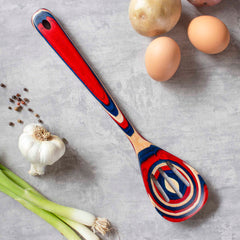 A vibrant Baltique® Old Glory Collection Slotted Spoon, 12-1/2" by Totally Bamboo, featuring red, blue, and natural wood patterns, rests on a gray surface. Around it are three eggs, a garlic bulb, a potato, scattered peppercorns, and green onions.