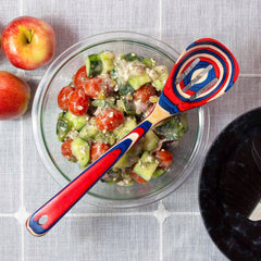 A bowl of salad with cucumbers, cherry tomatoes, and feta cheese sits on a tablecloth next to two apples. Resting on top of the salad is a colorful slotted spoon from the Totally Bamboo Baltique® Old Glory Collection. A black plate is partially visible nearby.