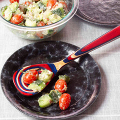 A vibrant cucumber and cherry tomato salad with a creamy dressing is served in a clear bowl. On a black plate, a serving of the salad is presented with the Baltique® Old Glory Collection Slotted Spoon by Totally Bamboo, showcasing red, blue, and natural wood tones. The scene is set against a light tablecloth backdrop.