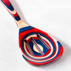 The Baltique® Old Glory Collection Slotted Spoon, 12-1/2" by Totally Bamboo showcases a vibrant swirling pattern in red, blue, and white on colored birch. It rests on a light gray surface, offering a visually striking effect that combines functionality with decoration.