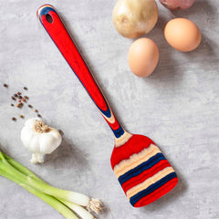 A Baltique® Old Glory Collection Spatula, 12-1/2" by Totally Bamboo, lies on a gray surface surrounded by two eggs, three potatoes, a garlic bulb, two green onions, and sprinkled black peppercorns.