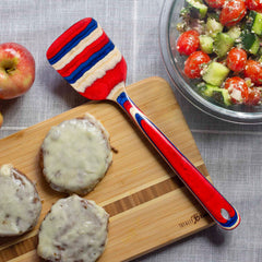 A 12-1/2" Baltique® Old Glory Collection Spatula by Totally Bamboo sits on a wooden cutting board with three cheese-covered patties. Nearby is a bowl of salad featuring cherry tomatoes, cucumbers, and feta cheese, while an apple rests on a light gray tablecloth.