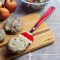 Cheeseburgers with melted cheese are placed on a wooden cutting board, surrounded by an apples and salad bowl backdrop. Adding a touch of patriotic flair to this classic scene is the vibrant Baltique® Old Glory Collection Spatula, 12-1/2" from Totally Bamboo, featuring stars and stripes.