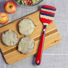 Three cheeseburgers rest on a wooden cutting board, accompanied by apples and a bowl of salad, with a vibrant Totally Bamboo Baltique® Old Glory Collection Spatula, 12-1/2", lying beside them on a gray cloth background.