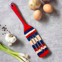 A 13-inch multicolored wooden spurtle from the Baltique® Old Glory Collection by Totally Bamboo, showcasing vibrant red, blue, and natural wood tones, rests on a gray surface. In close proximity are two brown eggs, a potato, a garlic bulb, green onions, and scattered black peppercorns—this versatile kitchen tool is poised for any culinary adventure.