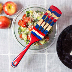 A vibrant spurtle from Totally Bamboo's Baltique® Old Glory Collection, featuring a red, blue, and beige pattern, rests on a bowl of chopped vegetables. Two apples are placed nearby on a gray tablecloth, highlighting the flair of this versatile 13" kitchen tool in any culinary setting.