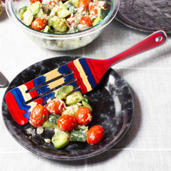 A black plate featuring a vibrant spatula, such as the Baltique® Old Glory Collection Spurtle, 13" by Totally Bamboo in white and blue, holds a salad of cucumber, cherry tomatoes, and cheese. Another bowl of salad sits in the background on a white tablecloth.