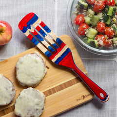 Part of the Baltique® Old Glory Collection from Totally Bamboo, a vibrant red, blue, and beige spurtle with an eye-catching design rests on a cutting board with three burger patties topped with melted cheese. Nearby, a salad and apple sit casually on the tablecloth.