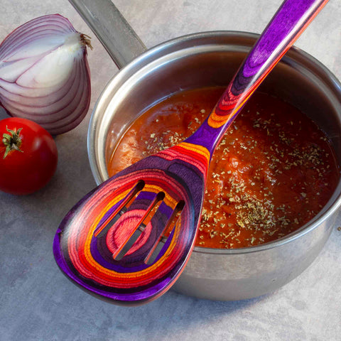 A vibrant Baltique® Waikiki Collection Slotted Spoon, 12-1/2" by Totally Bamboo rests on a pot filled with tomato sauce, sprinkled with herbs. A red onion and a tomato sit on the table beside the pot, enhancing the colorful vibes of this culinary scene.