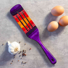 A vibrant Baltique® Waikiki Collection Spurtle, 13" by Totally Bamboo, adorned with patterns in purple, red, yellow, and black rests on a gray surface. This colorful kitchen tool lies alongside three brown eggs, a garlic bulb, and scattered peppercorns and is safe for non-stick cookware.