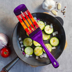 A vibrant kitchen utensil, the Baltique® Waikiki Collection Spurtle by Totally Bamboo, rests on a frying pan filled with sliced zucchini and chopped onions sprinkled with herbs. Surrounding the pan are a red onion, tomato, garlic bulb, cloves, and peppercorns—all set on a gray countertop. It's perfectly safe for non-stick cookware.