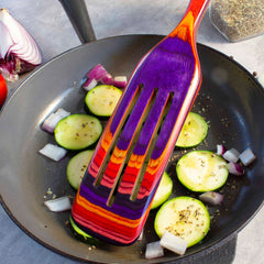 A vibrant Baltique® Waikiki Collection Spurtle, 13" by Totally Bamboo, stirs slices of zucchini and chopped red onions in a black frying pan. The vegetables are seasoned with herbs. Safe for non-stick cookware, this colorful kitchen tool adds flair as a quarter of a red onion and a spice jar sit in the background.