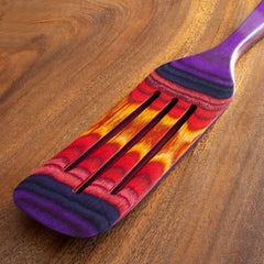 The Totally Bamboo Baltique® Waikiki Collection Spurtle, a 13" kitchen tool adorned with a vibrant pattern of red, orange, purple, and black hues, is placed on a wooden surface. Its sleek and ergonomic design is ideal for non-stick cookware and includes three slots on its head.