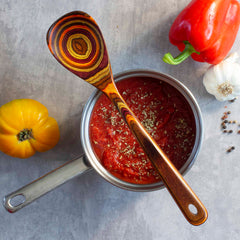 A saucepan filled with tomato sauce, sprinkled with herbs, is placed on a gray surface. Resting on the pan is a Totally Bamboo Baltique® Poconos Collection Cooking Spoon, 12-1/2", featuring a patterned handle. Nearby lies a red bell pepper, a yellow tomato, garlic, and peppercorns, creating an array of autumnal colors.