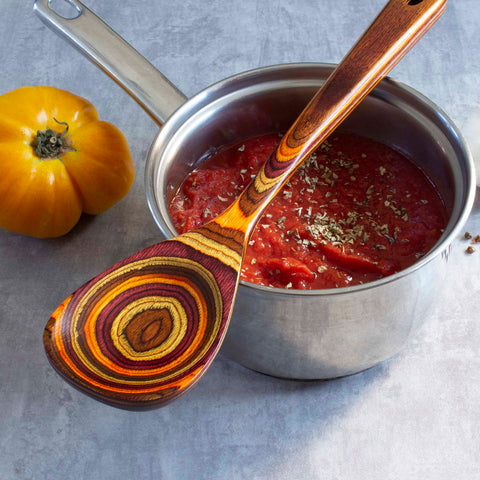 A stainless steel pot filled with tomato sauce and garnished with herbs sits on a gray surface, showcasing rich autumnal hues. The vibrant Totally Bamboo Baltique® Poconos Collection Cooking Spoon, 12-1/2", rests on the pot's non-stick safe edge. A yellow tomato lies next to the pot.