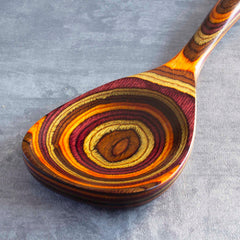 The Totally Bamboo Baltique® Poconos Collection Cooking Spoon, measuring 12-1/2 inches, showcases a smooth and polished finish with vibrant concentric rings in autumn-inspired colors such as brown, yellow, and red, set against a textured gray backdrop. Its non-stick safe design makes it both functional and artistically striking.