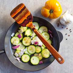 A pan filled with sliced zucchini, chopped onions, and seasonings rests on a gray surface. Inside the pan is a wooden Totally Bamboo Baltique® Poconos Collection Spatula, 12-1/2". Nearby are a yellow tomato, a garlic bulb, and scattered peppercorns that reflect the rich autumnal hues of colored birch.