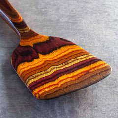 A close-up of the Totally Bamboo Baltique® Poconos Collection Spatula, 12-1/2", highlights its autumnal palette with a vibrant, multicolored striped design in shades of orange, brown, and yellow. Made from colored birch, it rests gracefully on a textured gray surface, producing a striking marbled effect.