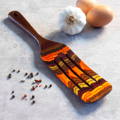 A vibrant 13-inch wooden spatula from the Baltique® Poconos Collection by Totally Bamboo, showcasing a versatile autumnal palette of brown, orange, and yellow, rests on a gray surface. Nearby, two eggs, a head of garlic, and scattered peppercorns complete the charming scene.