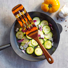 Sliced zucchini and diced onions sizzle in a frying pan on a gray surface, with an elegantly placed Baltique® Poconos Collection Spurtle, 13" by Totally Bamboo resting on top. Nearby are a yellow tomato, a garlic bulb, and black peppercorns—capturing the essence of autumnal colors.