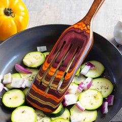 A vibrant, layered wooden spatula from Totally Bamboo's Baltique® Poconos Collection Spurtle, 13", stirs sliced zucchini and purple onions in a black pan. A yellow tomato and a garlic bulb, showcasing autumnal colors, are placed nearby on a light gray countertop.