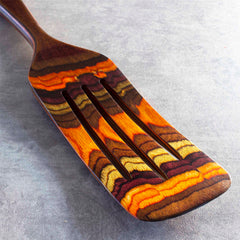 A wooden fork from the Baltique® Poconos Collection by Totally Bamboo, showcasing a vibrant, layered design in autumn-inspired shades of orange, brown, and yellow. This versatile kitchen tool features three prongs and rests on a textured gray surface, exuding an artistic charm similar to the renowned 13-inch spurtle from the same collection.