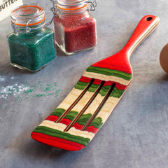 A multicolored spatula with red, green, and beige stripes from the Totally Bamboo Baltique® North Pole Collection Spurtle, 13" rests on a gray surface. In the background, glass jars filled with festive Christmas-colored sugar and a partially visible egg enhance the kitchen's cheerful ambiance.