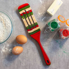 A festive 13" spurtle from the Totally Bamboo Baltique® North Pole Collection, featuring red, green, and natural wood stripes, rests on a countertop sprinkled with flour, two eggs, butter, and jars of Christmas-colored sprinkles. This enchanting kitchen tool creates the perfect backdrop for a delightful baking scene.