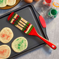 A beautifully crafted spurtle from the Baltique® North Pole Collection by Totally Bamboo rests on a baking tray of sugar cookies adorned with red and green sprinkles. Nearby, eggs and jars of Christmas-colored sugar enhance the festive atmosphere on the gray countertop, completing this charming kitchen scene.